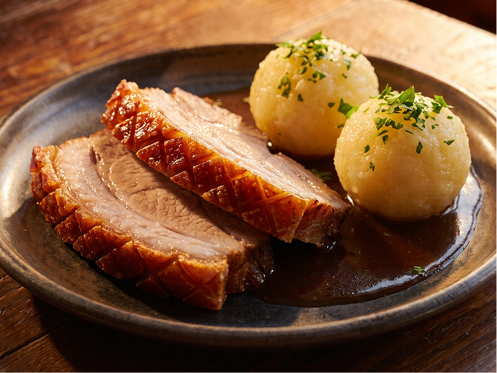 Zwei Scheiben bayerischer Schweinebraten mit knuspriger Kruste, serviert mit zwei Kartoffelknödeln und dunkler Bratensauce auf einem rustikalen Teller.