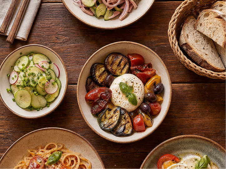 Flache Draufsicht auf einen Holztisch mit verschiedenen italienischen Gerichten, darunter eine große Schale Burrata mit gegrillten Auberginen, Kirschtomaten und Oliven, ein Gurkensalat, Pasta und ein Brotkorb.