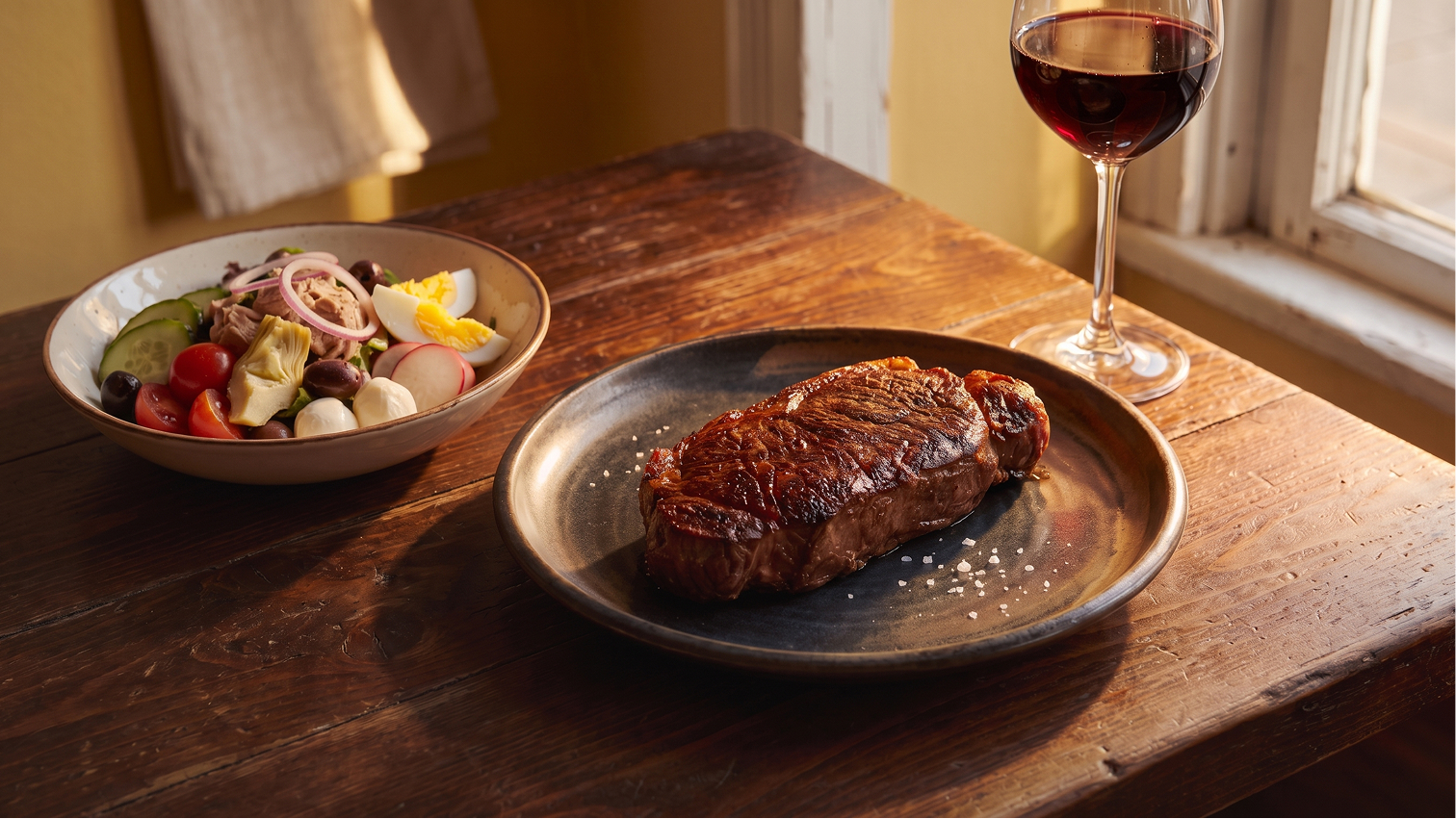 Ein saftig gebratenes Rindersteak mit grobem Salz auf einem dunklen Teller, serviert mit einem bunten Beilagensalat und einem Glas Rotwein auf einem rustikalen Holztisch.